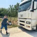 Eine Person mit Mütze, Sonnenbrille, schwarzem Hemd und blauen Shorts geht auf einem unbefestigten Weg neben einem großen weißen MAN-LKW und gestikuliert mit einem Lächeln. Im Hintergrund sind Bäume und trockenes Gras an einem sonnigen Tag zu sehen.