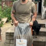 Ein Mann mit Tätowierungen hält eine große Tüte Hundefutter mit der Aufschrift Tierheim Tierfutter in der Hand. Er steht draußen in der Nähe eines Gartens, in dem zwei schwarze Hunde leben.