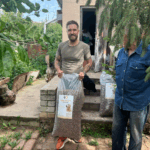 Ein Mann steht im Freien, hält eine große Tüte mit Hundefutter in der Hand und lächelt in die Kamera. Eine andere Person steht in der Nähe, teilweise außerhalb des Bildes. Im Hintergrund sind Grünflächen und ein Haus zu sehen.
