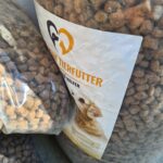 Zwei große Plastiktüten, gefüllt mit Trockenfutter für Haustiere. Der vordere Beutel ist mit einem Etikett versehen, das einen Hund und einen Teil des Textes in deutscher Sprache zeigt, wobei das Trockenfutter durch die durchsichtige Verpackung sichtbar ist.