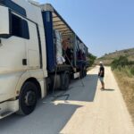 Ein Mann steht auf einer unbefestigten Straße neben einem großen weißen Lastwagen, dessen Anhänger an der Seite geöffnet ist, so dass die gestapelte Ladung im Inneren sichtbar wird. Die sonnige Landschaft umfasst grasbewachsene Felder und einen Hügel im Hintergrund.