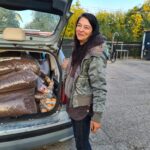 Eine Frau steht neben einem offenen Kofferraum, der mit großen Säcken mit Tierfutter gefüllt ist. Sie ist im Freien, trägt eine Jacke und lächelt in die Kamera. Im Hintergrund sind Bäume und eine Tankstelle zu sehen.