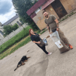 Eine Frau und ein Mann stehen draußen in der Nähe einer großen Tüte mit Hundefutter; die Frau hält einen Behälter, und ein schwarz-brauner Hund liegt in der Nähe auf dem Boden. Im Hintergrund sind Gebäude und Grünflächen zu sehen.