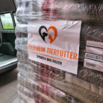 Eine in Plastik eingewickelte Palette mit Säcken mit Tierfutter, mit der Aufschrift "Tierheim Tierfutter Spenden und Helfen" und dem Bild einer Katze und eines Hundes. Die Palette steht neben einem offenen Fahrzeug.