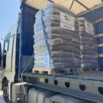 Ein Lastwagen mit geöffneter Seite zeigt mehrere gestapelte Paletten mit verpackten Waren, die in Plastik eingewickelt sind. Die Verpackungen sind mit Etiketten versehen, auf denen ein Logo mit einem Herz und Text zu sehen ist, und der Lkw ist an einem sonnigen Tag im Freien geparkt.