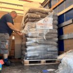 Eine Person in kurzen Hosen und blauem Hemd entlädt dicht verpackte Säcke, die auf einer Holzpalette gestapelt sind, aus einem Lastwagen. Im Inneren des Lastwagens sind ebenfalls große Kisten und in Plastik verpackte Gegenstände zu sehen.