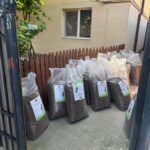 Große Säcke mit Tierfutter sind vor einem roten Holzzaun vor einem Haus aufrecht gestapelt. Jede Tüte hat ein Etikett mit Tierbildern und Text. Der Bereich liegt im Schatten der nahen Bäume.