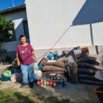 Eine Frau steht draußen vor einem Haus neben Säcken mit Tierfutter, Konserven und Vorräten, die sich auf dem Boden stapeln. Ein schwarzes Auto und eine Schubkarre stehen in der Nähe, das Sonnenlicht wirft scharfe Schatten.