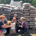 Vier Personen knien im Freien mit Hunden vor gestapelten Tüten mit Tierfutter. Auf einem Schild oben steht VIELEN DANK! mit einem Herz. Im Hintergrund sind Bäume und Grünpflanzen zu sehen.