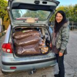 Eine Frau mit langen dunklen Haaren und einer grünen Jacke steht lächelnd neben einem offenen Kofferraum, der mit großen Tüten mit braunem Tierfutter gefüllt ist. Es ist Tag, und die Szene spielt sich draußen in einer Einfahrt ab.