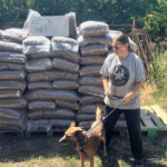 Eine Frau, die einen braunen Hund an der Leine hält, steht vor gestapelten Tüten, auf denen ein Schild mit der Aufschrift Vielen Dank! und ein Herz zu sehen ist. Im Hintergrund sind Bäume und Holzpaletten zu sehen, an einem sonnigen Tag.