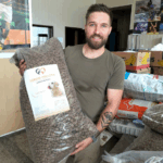 Ein bärtiger Mann mit einer Tätowierung auf dem linken Unterarm lächelt und hält eine große Tüte mit Hundefutter in der Hand. Hinter ihm stapeln sich mehrere andere Tüten mit Tierfutter in einem Raum mit Kartons und Postern an den Wänden.