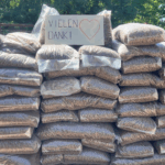 Ein großer Stapel mit Pellets gefüllter Plastiksäcke ist im Freien aufgestellt; obenauf ist ein Schild mit der Aufschrift VIELEN DANK! und einem roten Herz zu sehen, auf dem in deutscher Sprache gedankt wird. Im Hintergrund sind Bäume und Sonnenlicht zu sehen.