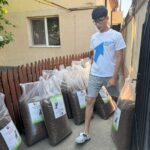 Ein junger Mann in weißem T-Shirt und kurzen Hosen steht im Freien neben mehreren großen durchsichtigen Säcken mit Tierfutter, die an einem Holzzaun gestapelt sind. In der Nähe gibt es ein Gebäude mit einem Fenster und Bäumen.