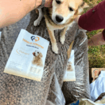 Eine Person streichelt einen hellbraunen und weißen Hund, der auf zwei großen Säcken mit Hundefutter mit der Aufschrift Tierheim Tierfutter im Freien auf einer Wiese steht. Der Arm einer anderen Person ist neben den Vorräten zu sehen.