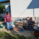 Eine Frau steht draußen an einer Mauer mit Tüten mit Tierfutter, Konserven und Vorräten, die im Sonnenlicht auf dem Boden gestapelt sind. Auch eine Schubkarre und ein Auto sind in der Nähe zu sehen.