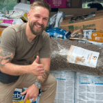 Ein lächelnder Mann mit Bart und Tattoos zeigt den Daumen nach oben vor Stapeln von großen Tüten und Kartons mit Tierfutter und deutet damit auf eine Lieferung oder Spende an.