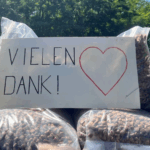 Ein handgeschriebenes Schild mit der Aufschrift VIELEN DANK! und einem roten Herz befindet sich auf mehreren großen, mit braunen Pellets gefüllten Plastiksäcken, die im Freien stehen, während im Hintergrund Bäume zu sehen sind.