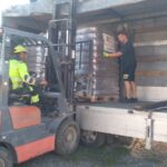 Ein Gabelstaplerfahrer in Warnkleidung lädt in Schrumpffolie verpackte Paletten auf einen Lkw, während ein Mann in schwarzen Shorts und T-Shirt die Paletten in den Lkw führt.