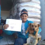 Ein Mann in blauer Jacke und Mütze sitzt vor gestapelten Tüten, hält einen kleinen braunen Hund und ein handgeschriebenes Schild mit Danke und einem Herz in der Hand.