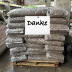 Ein Stapel mit braunen Pellets gefüllter Plastiksäcke liegt auf einer Holzpalette in einer Industrieumgebung. An dem Stapel ist ein weißes Schild mit dem Wort "Danke" angebracht.
