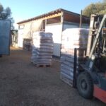 Ein Arbeiter bedient einen Gabelstapler, um eine Palette mit gestapelten, verpackten Säcken zu bewegen, während sich in der Nähe weitere Paletten und ein Lieferwagen mit offenen Türen in einem Außenlager befinden.