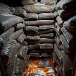 In einem Lagerraum stapeln sich große, durchsichtige Plastiktüten, die mit braunen Pellets gefüllt sind, an den Wänden. Leuchtend orangefarbene und silberne Säcke liegen bei gedämpfter Beleuchtung auf dem Boden verstreut.