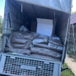 Große, mit braunen Pellets gefüllte Plastiksäcke stapeln sich im hinteren Teil eines offenen Lastwagens, der in der Nähe eines Zauns und eines Gebäudes mit lila Dach geparkt ist.