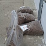 Große Plastiktüten, gefüllt mit braunem Tierfutter, sind auf einer Betonfläche neben einer weißen Wand und einem Abflussrohr gestapelt. Im Hintergrund ist ein Hund zu sehen, der davonläuft.