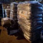 In einer schwach beleuchteten Scheune lagern vier große, in Plastik eingewickelte Paletten mit Säcken voller Tierfutter, die mit "Tierheim Tierfutter" beschriftet sind. Eine Schubkarre mit Tierfutter steht neben den Paletten. Ein deutscher Text überlagert das Bild.