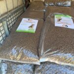 Große durchsichtige Plastiktüten, gefüllt mit Kroketten oder Trockenfutter, sind auf einer Palette gestapelt. Jeder Beutel ist mit einem Etikett versehen, auf dem eine Katze und Produktinformationen abgebildet sind. Der Schauplatz scheint ein Lager- oder Versorgungsbereich zu sein.