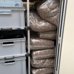 Große durchsichtige Plastiktüten, gefüllt mit braunem Tierfutter, stapeln sich auf Regalen in einem Lagerraum neben Plastikbehältern mit Schubladen.