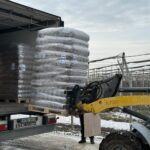 Ein Gabelstapler entlädt eine große, in Plastik verpackte Palette von einem Lastwagen auf einem verschneiten Gelände in der Nähe eines eingezäunten Bereichs mit kahlen Bäumen im Hintergrund.