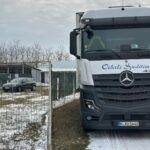 Ein großer Mercedes-Benz-Lkw ist neben einem Maschendrahtzaun auf einem schneebedeckten Gelände geparkt, in der Nähe eines grauen Gebäudes und eines geparkten Autos, mit blattlosen Bäumen und einem bewölkten Himmel im Hintergrund.