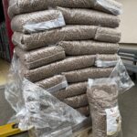 Große Stapel durchsichtiger Plastiksäcke, die mit braunen Pellets gefüllt sind, stapeln sich auf einer Palette im Freien. Ein Sack ist an der Vorderseite geöffnet und zeigt den Inhalt und ein Produktetikett. Einige Plastikverpackungen sind lose um die Palette herum.