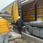 Drei Personen entladen große Säcke mit Waren, möglicherweise Lebensmittel oder Vorräte, aus einem gelben Sattelschlepper, der am Straßenrand geparkt ist. Die Männer tragen warme Kleidung, und die Seitenplane des Lkw ist geöffnet.