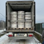 Ein Kühltransporter mit geöffneten Hecktüren zeigt Stapel von großen, verpackten, runden Paketen im Inneren. Der Lkw ist auf einer verschneiten Straße geparkt, mit einer Umzäunung und einer Person, die in der Nähe steht.