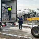 Ein Mann in einer gelben Weste steht in einem Lastwagen und richtet die gestapelten Säcke aus, während ein anderer Mann in der Nähe einen Gabelstapler auf einem verschneiten Weg im Freien neben einem Gewächshaus bedient.