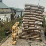 Ein Stapel mit Pellets gefüllter Plastiksäcke ist auf einer Holzpalette im Freien in der Nähe eines rustikalen Gebäudes aufgestapelt, in der Nähe befindet sich eine leere Palette und ein grasbewachsener Boden. Im Hintergrund sind ein bedeckter Himmel und Stromleitungen zu sehen.