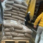 Ein hoher Stapel großer Plastiksäcke, gefüllt mit etwas, das wie Bohnen oder Getreide aussieht, liegt auf einer Holzpalette neben einem offenen gelben Lastwagen. Mehrere Menschen in Freizeitkleidung stehen in der Nähe, und der Himmel ist bedeckt.