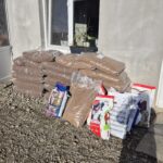 Große Stapel von Plastiktüten mit Tierfutter und mehrere bunte Tüten mit Hunde- und Katzentrockenfutter stapeln sich vor einem Gebäude mit einem Fenster und einer Tür auf einer Kiesfläche.