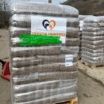 Eine große Palette mit Säcken mit Tierfutter, die in Plastik eingewickelt sind, steht im Freien auf einer unbefestigten Fläche. An der Vorderseite des Stapels ist ein Schild mit Tiermotiven und Text angebracht. Im Hintergrund sind weitere Paletten zu sehen.