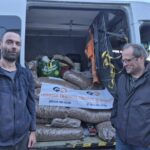 Zwei Männer stehen neben einem offenen Lieferwagen, der mit großen Säcken mit Tierfutter und -zubehör gefüllt ist. Auf einem Transparent am Wagen steht TIERHEIM TIERFUTTER SPENDEN UND HELFEN auf Deutsch.