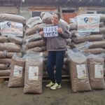 Eine lächelnde Person hält ein "DANKE!"-Schild in die Höhe, während sie vor einem Gebäude vor großen Stapeln von Tierfuttertüten steht, von denen einige mit "Streethearts Together" beschriftet sind.