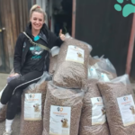 Eine lächelnde Frau sitzt auf großen Tüten mit Hundefutter, die vor einem Gebäude gestapelt sind, und zeigt den Daumen nach oben. Die Tüten sind mit Etiketten mit Hundebildern versehen, und in der oberen rechten Ecke ist ein blaugrüner Pfotenabdruck abgebildet.