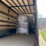 Ein großer, metallischer Wassertank mit einem Etikett steht in einem leeren, offenen Lkw, der auf einer Landstraße geparkt ist. Auf dem Boden des Lastwagens sind Gurte und Geräte zu sehen.