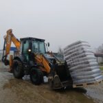 Ein gelber CASE-Baggerlader hebt eine große Palette mit Säcken auf einer schlammigen Baustelle, mit einigen Gebäuden und blattlosen Bäumen im Hintergrund unter einem bedeckten Himmel.