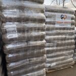 Im Freien sind große Stapel mit Paletten verpackter Tiereinstreu oder Futtersäcke zu sehen. Auf einer Palette ist ein Schild mit der Aufschrift Tierheim Tierfutter zu sehen, das darauf hinweist, dass es sich um Tierheimbedarf handelt. Im Hintergrund sind blattlose Bäume zu sehen.