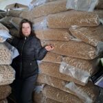 Eine Frau in einer schwarzen Jacke steht in einem Lagerraum neben großen Stapeln versiegelter Plastiktüten, die mit braunem Tierfutter gefüllt sind.