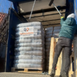 Eine Person in grüner Jacke reinigt mit einem Besen die Oberseite eines offenen Transportcontainers, der mit gestapelten Säcken mit der Aufschrift Tierheim Tierfutter gefüllt ist. Das Sonnenlicht wirft Schatten auf die Kisten und Paletten im Inneren.